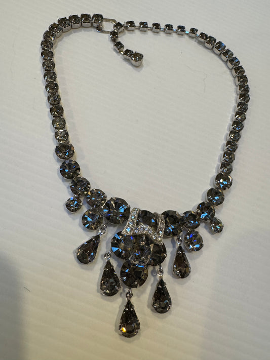Channel vintage sophistication with this showstopping 1950s Weiss rhinestone necklace, designed in a dramatic bib style. Crafted with dazzling smoky grey crystals and suspended teardrop dangles, this piece is the epitome of mid-century evening elegance.