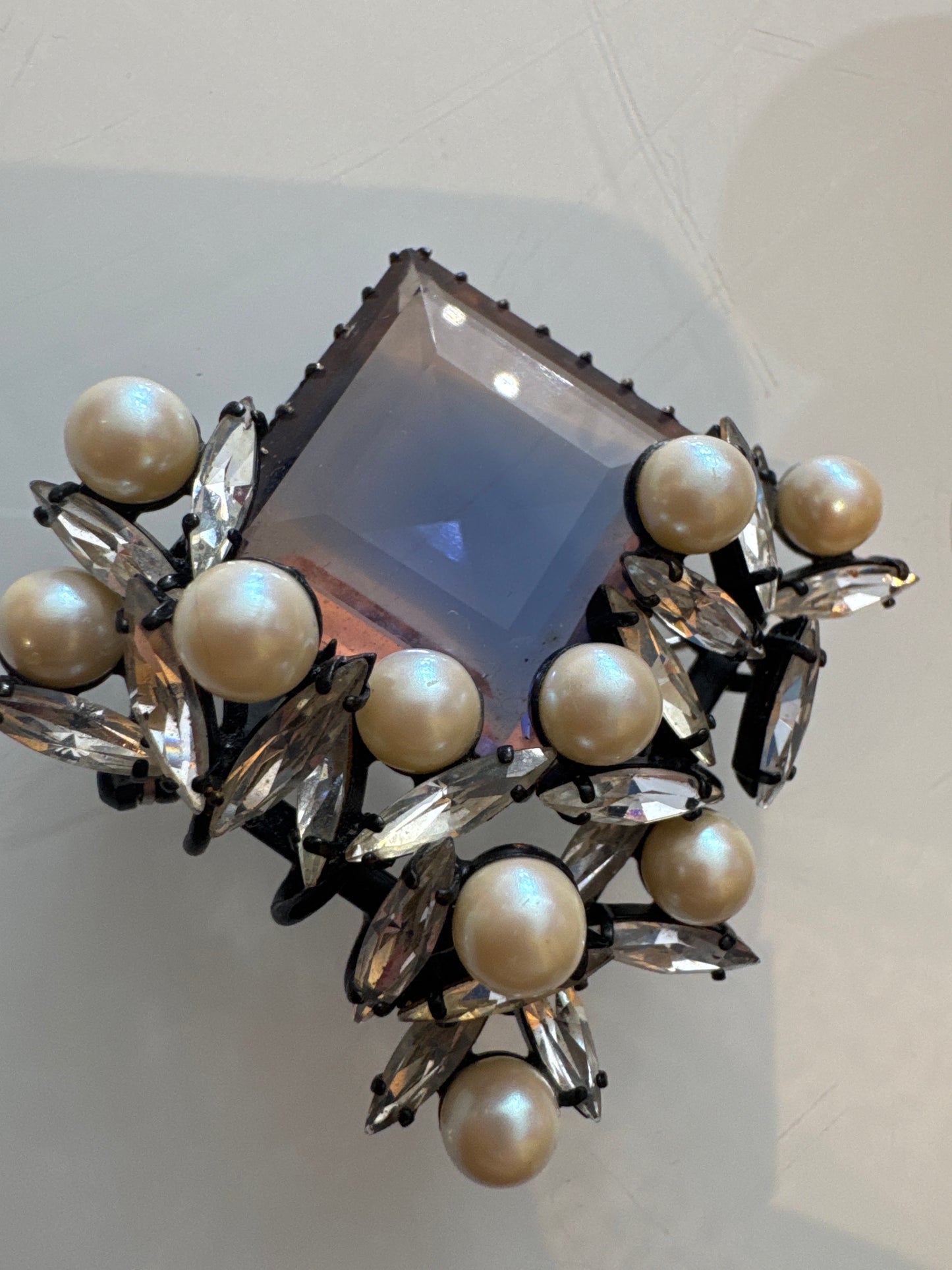 Sculptural mid-century couture brooch featuring a faceted lavender glass center stone, framed by faux pearls and clear navette rhinestones in black japanned metal. Atelier-made, circa 1950s–60s. Architectural, dramatic, and exquisitely constructed.