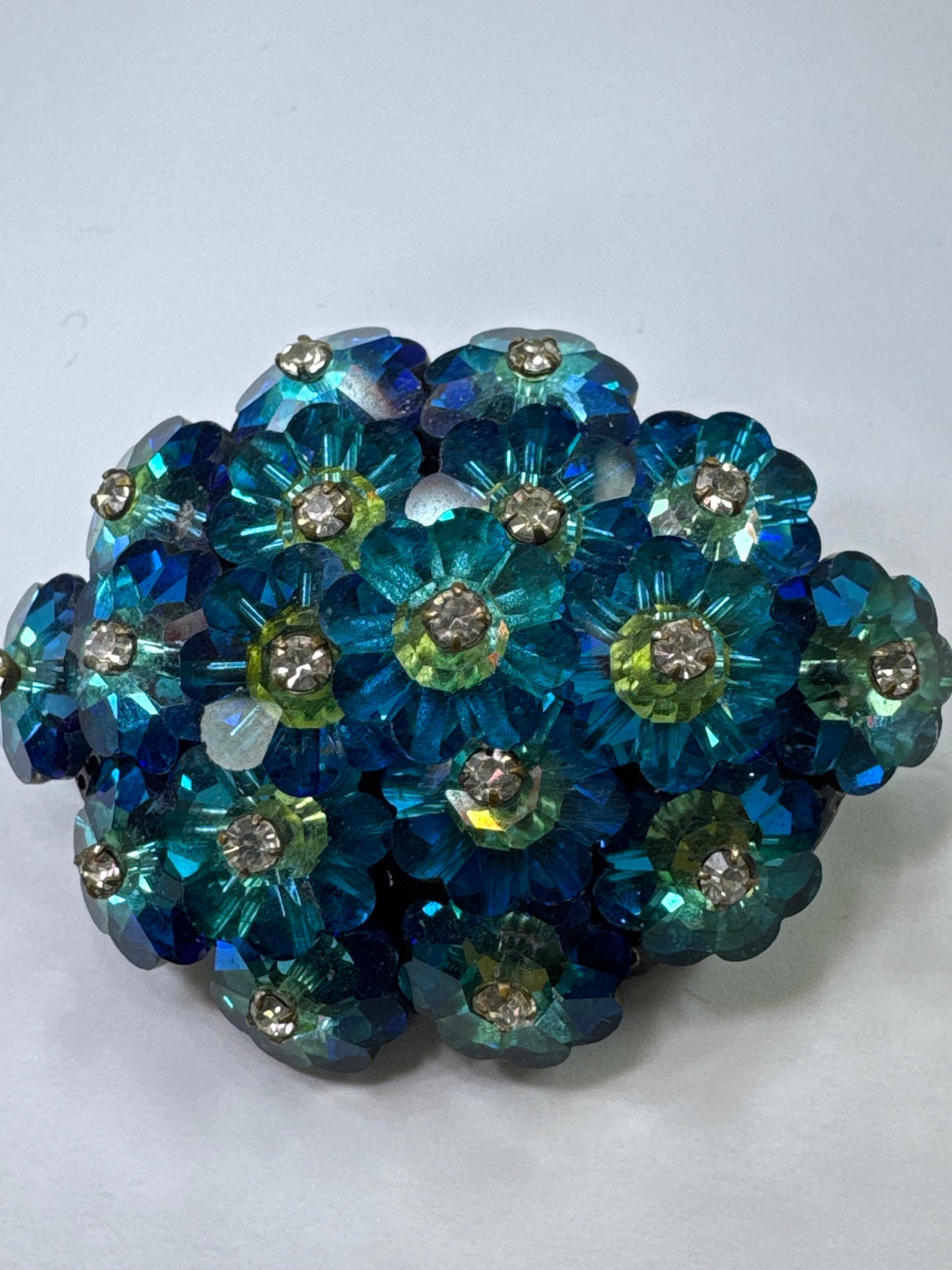 A striking vintage brooch composed of layered, faceted crystal florets in rich shades of teal, emerald, and deep ocean blue. Each petal-like stone is cut to catch the light from every angle, creating a dimensional, almost floral cluster effect that feels
