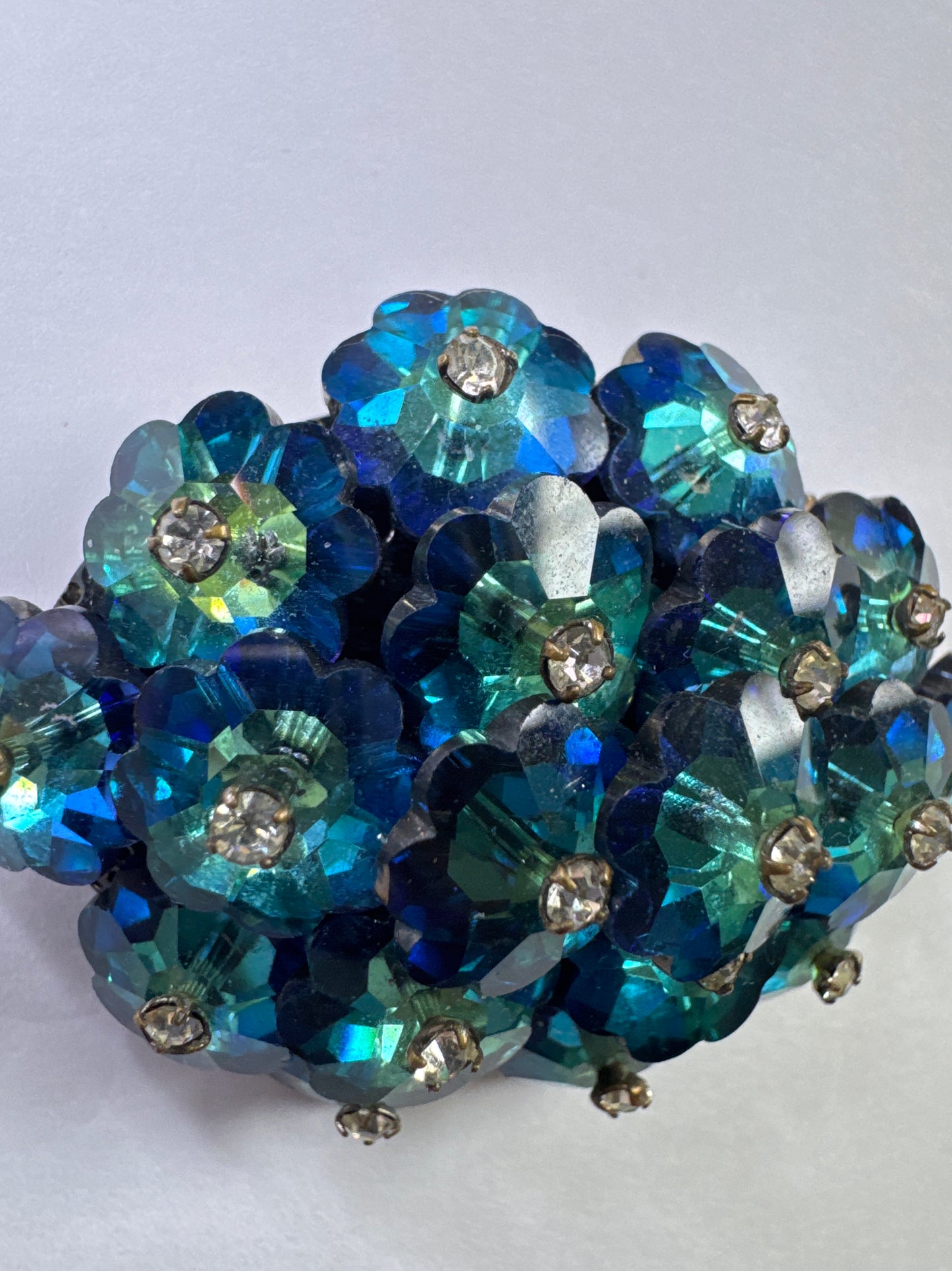 A striking vintage brooch composed of layered, faceted crystal florets in rich shades of teal, emerald, and deep ocean blue. Each petal-like stone is cut to catch the light from every angle, creating a dimensional, almost floral cluster effect that feels